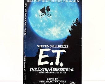 May include: A paperback book titled "E.T. The Extra-Terrestrial" by William Kotzwinkle. The cover features a silhouette of a person and E.T. on a bicycle in front of a blue moon. The book is a Special Book Club Edition.