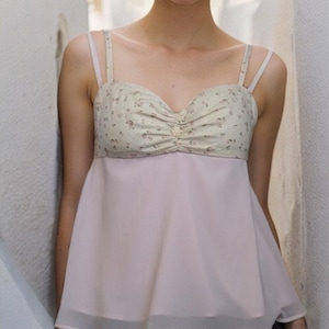 May include: A young woman wearing a pink and white floral print top with a sheer overlay. The top has thin straps and a sweetheart neckline. She is wearing black jeans.