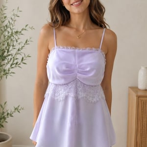 May include: A light purple camisole top with spaghetti straps and a square neckline. The top features a gathered bodice and a white lace trim along the neckline and hem. The model is wearing blue jeans.