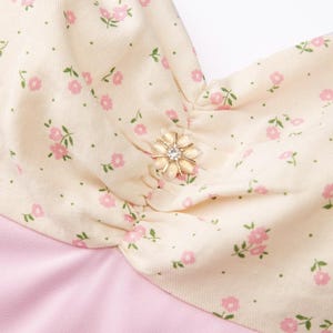 May include: A close-up of a cream-colored fabric with a small pink floral pattern. The fabric has a gathered detail and a small gold button with a clear rhinestone in the center. The fabric is layered over a solid pink fabric.