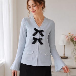 May include: A light blue button-down top with black bow accents and two front pockets. The top features a V-neckline with lace trim and flared sleeves with lace detailing. The model is wearing a black skirt.