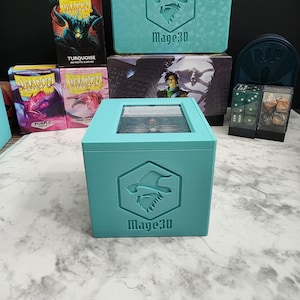 May include: A turquoise blue cube-shaped box with a clear plastic window on the front. The box has a logo on the front that says "Mage3D" and a stylized image of a wizard with a beard and a hat. The box is sitting on a white marble surface.