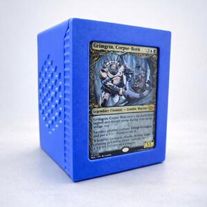 May include: A blue cube-shaped card holder with a Magic: The Gathering card inside. The card features the character Grimgrin, Corpse-Born, a Zombie Warrior. The holder has a perforated side.