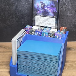 Op de afbeelding: Een blauwe plastic kaartenhouder met een grote stapel blauw gesleevde ruilkaarten, een zijstapel kaarten en een rij veelkleurige dobbelstenen. Een enkele ruilkaart met de titel Zul Ashur, Lich Lord staat rechtop in de achterste gleuf.