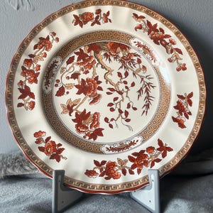 Op de afbeelding: Een decoratieve keramische plaat met een crèmekleurige achtergrond en een gedetailleerd bloemmotief in tinten rood, bruin en goud. De plaat heeft een geschulpte rand en een patroonrand, die een klassieke esthetiek laat zien. Diameter: ongeveer 25 cm.