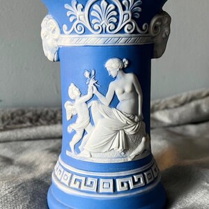 May include: Blue and white ceramic vase with classical relief figures. The vase features a detailed design of a woman and cherub, with decorative elements around the top and base. The vase is approximately 6 inches tall.