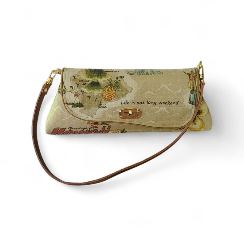 Puede incluir: Bolso de hombro beige con correa marr&oacute;n y herrajes dorados. El bolso presenta un mapa de Haw&aacute;i con ilustraciones de una pi&ntilde;a y personas. El texto incluye "Aloha" y "Life is one long weekend."