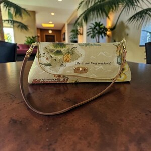 Puede incluir: Bolso de mano beige con un mapa de Haw&aacute;i, una pi&ntilde;a y las palabras "Aloha" y "Life is one long weekend". El bolso tiene una correa marr&oacute;n y herrajes dorados. "Greetings from Hawaii" est&aacute; impreso en la parte inferior.