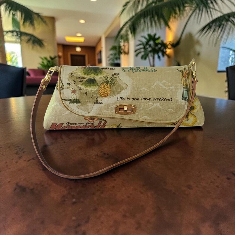 Puede incluir: Bolso de mano beige con un mapa de Haw&aacute;i, una pi&ntilde;a y las palabras "Aloha" y "Life is one long weekend". El bolso tiene una correa marr&oacute;n y herrajes dorados. "Greetings from Hawaii" est&aacute; impreso en la parte inferior.