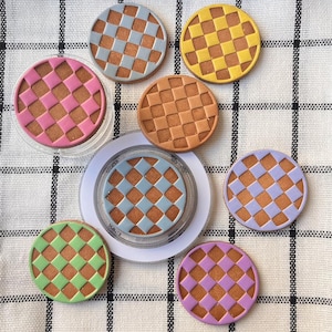 Può includere: Una collezione di sottobicchieri rotondi e colorati con un motivo a scacchiera. I sottobicchieri presentano vari colori, tra cui rosa, blu, giallo, arancione, verde e viola. Alcuni sottobicchieri sono in supporti di plastica trasparente.