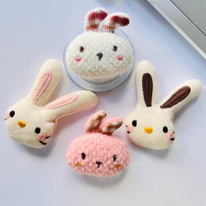 May include: Four plush bunny-shaped accessories in white and pink. The bunnies have embroidered facial features, including black eyes, pink cheeks, and yellow noses. Two bunnies have plaid ears. One bunny is attached to a clear circular base.