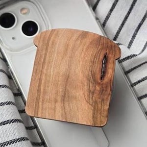 May include: A wooden phone grip shaped like a slice of bread. The grip is made of light brown wood with a natural grain pattern and a dark vertical streak. The phone is in a clear case, resting on a striped fabric.