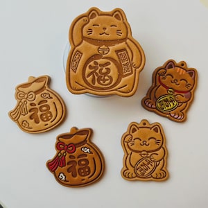 May include: A collection of brown Maneki Neko lucky cat charms and fortune bag charms. The charms feature the Japanese kanji for fortune, and are in various poses, including waving and holding coins. The charms are made of a light brown material.