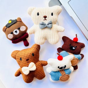 Könnte beinhalten: Fünf Plüsch-Teddybär-Ornamente in verschiedenen Farben und Stilen. Einer ist cremefarben mit einer grauen Fliege, ein anderer ist braun mit einer weißen Fliege, und ein dritter trägt einen Hut und einen Schal. Zwei weitere haben Kirschen.