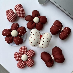 Puede incluir: Una variedad de accesorios para el cabello hechos a mano, de tela, con formas de flores y lazos. Los accesorios presentan una paleta de colores burdeos, blanco y rojo, con estampados de lunares y cuadros. Las flores tienen centros de botón blancos.