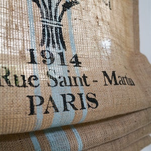 May include: A brown burlap roman shade with blue stripes and black text that reads "1914 Rue Saint-Martin PARIS".