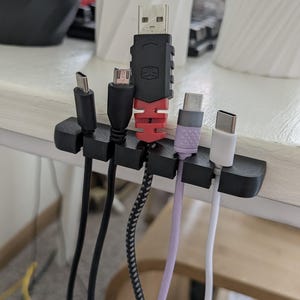 May include: A black cable organizer holding various USB cables. Cables include USB-C, micro-USB, and USB-A connectors in black, white, and purple. The organizer is on a light-colored surface.
