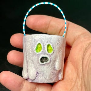 May include: A small, handcrafted ghost-shaped container with a white and purple body, green eyes, and a black mouth. It has a striped blue and white handle. The container is designed to resemble a ghost and is likely used for Halloween or spooky-themed decorations.