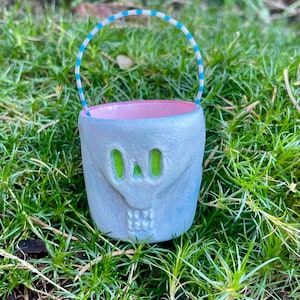 May include: A small, light blue skull-shaped bucket with a pink interior and a striped handle. The skull has green eye sockets and a toothy grin. The bucket is sitting in green grass.