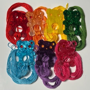 May include: A collection of colorful bear-shaped acrylic charms, each paired with a matching skein of embroidery floss. The charms are in shades of red, orange, yellow, green, blue, purple, and pink. The floss is neatly arranged beneath each charm.