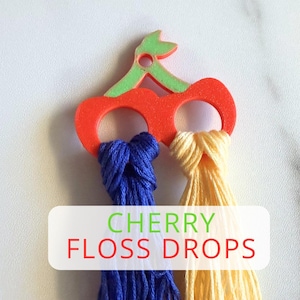 May include: A cherry-shaped floss drop holder in red and green, holding blue and yellow embroidery floss. The text "CHERRY FLOSS DROPS" is displayed on a white rectangle.