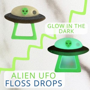 May include: Two alien UFO floss drops are shown, one in daylight and one glowing in the dark. The UFOs have a green alien face on top, a black rim, and a green stand. The text "GLOW IN THE DARK" and "ALIEN UFO FLOSS DROPS" are also visible.