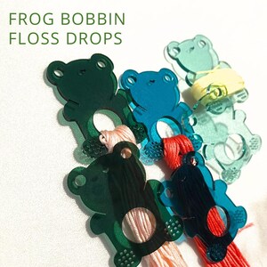May include: A collection of frog-shaped bobbins in shades of green and blue, holding colorful embroidery floss. The bobbins have a circular opening for the floss and a small hole for hanging. The text "FROG BOBBIN FLOSS DROPS" is in the upper left corner.