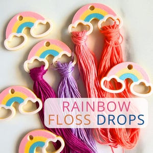May include: Rainbow floss drops with colorful embroidery thread. The image shows several rainbow-shaped floss drops in pink, yellow, and blue, each holding a bundle of thread in shades of pink, purple, and orange. The text "RAINBOW FLOSS DROPS" is also visible.
