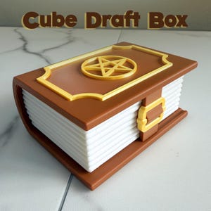 May include: A brown and gold Cube Draft Box, designed to resemble a book. The box features a gold pentagram on the cover and a gold buckle closure. The text "Cube Draft Box" is displayed above the box.