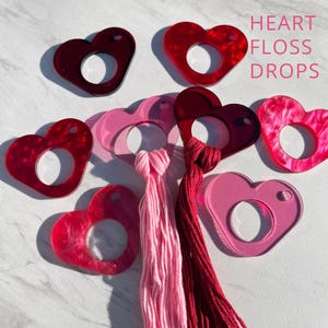 May include: A collection of heart-shaped floss drops in shades of red and pink. Some hearts are solid, others have a marbled effect. The text "HEART FLOSS DROPS" is visible. Pink and red floss is attached to some of the heart drops.