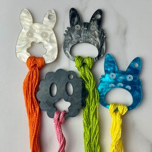 May include: A collection of whimsical bottle openers in the shape of cartoon characters. The openers are in white, gray, and blue, with orange, green, pink, and yellow tassels. The openers are made of acrylic.