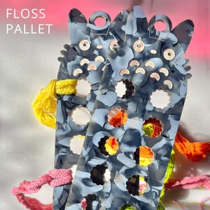 May include: Two blue and gray cat-shaped floss pallets with various colored floss and the text "FLOSS PALLET". The pallets have holes for holding floss and decorative cutouts. The floss colors include yellow, pink, and orange.