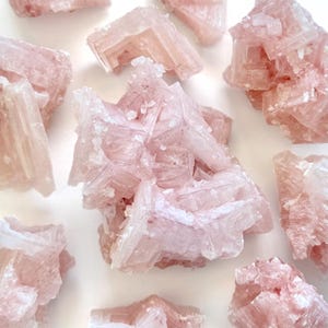 May include: A collection of raw, pale pink halite crystal clusters featuring cubic structures and translucent, jagged edges. The mineral specimens display varying shades of soft rose and white, with sharp geometric formations and a crystalline texture.