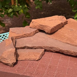 May include: A collection of irregular, flat, reddish-brown sandstone pieces stacked on top of a rectangular brick base. The stones feature rough, textured edges and natural, earthy tones, displaying a layered, sedimentary appearance throughout.