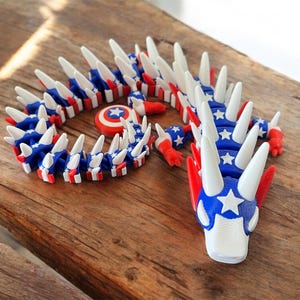 May include: A 3D-printed dragon toy in red, white, and blue, featuring a star-spangled design. The dragon has a segmented body and white spikes. A small shield with a star emblem is also present. The toy is displayed on a wooden surface.