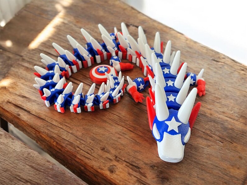 May include: A 3D-printed dragon toy in red, white, and blue, featuring a star-spangled design. The dragon has a segmented body and white spikes. A small shield with a star emblem is also present. The toy is displayed on a wooden surface.