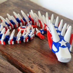 May include: A 3D-printed articulated dragon toy in red, white, and blue, with a patriotic theme. The dragon features a star-spangled head and body segments with alternating stripes. The toy is posed on a wooden surface.