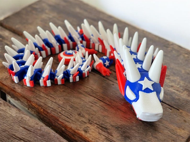 May include: A 3D-printed articulated dragon toy in red, white, and blue, with a patriotic theme. The dragon features a star-spangled head and body segments with alternating stripes. The toy is posed on a wooden surface.