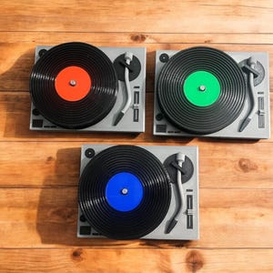 May include: Three gray record players with black vinyl records and colored center labels: red, green, and blue. The turntables are arranged on a wooden surface, showcasing the classic design of the record players.