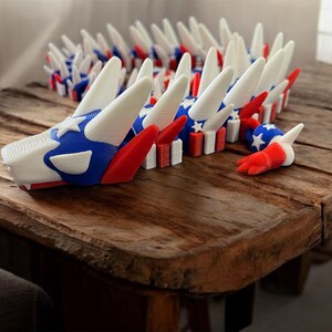 May include: A 3D-printed dragon sculpture in red, white, and blue, featuring a star-spangled head and tail. The dragon is segmented, with each section resembling a small flag. The sculpture is displayed on a rustic wooden table.