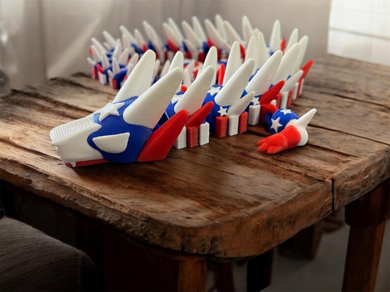 May include: A 3D-printed dragon sculpture in red, white, and blue, featuring a star-spangled head and tail. The dragon is segmented, with each section resembling a small flag. The sculpture is displayed on a rustic wooden table.