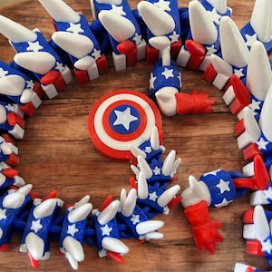 May include: A collection of 3D-printed toys in red, white, and blue, featuring star and stripe designs. The toys include a Captain America shield and a segmented, articulated toy. The toys are arranged on a wooden surface.