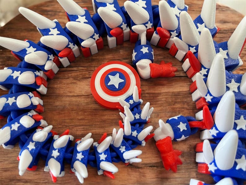 May include: A collection of 3D-printed toys in red, white, and blue, featuring star and stripe designs. The toys include a Captain America shield and a segmented, articulated toy. The toys are arranged on a wooden surface.