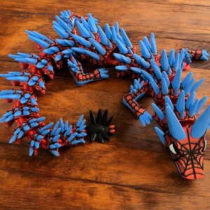 K&ouml;nnte beinhalten: Ein rotes und blaues, bewegliches Drachenspielzeug mit einem Spinnennetzdesign auf dem Kopf. Der Drache hat blaue Stacheln und ist aus Kunststoff. Ein schwarzes, stacheliges Zubeh&ouml;r liegt neben dem Drachen auf einer Holzoberfl&auml;che.