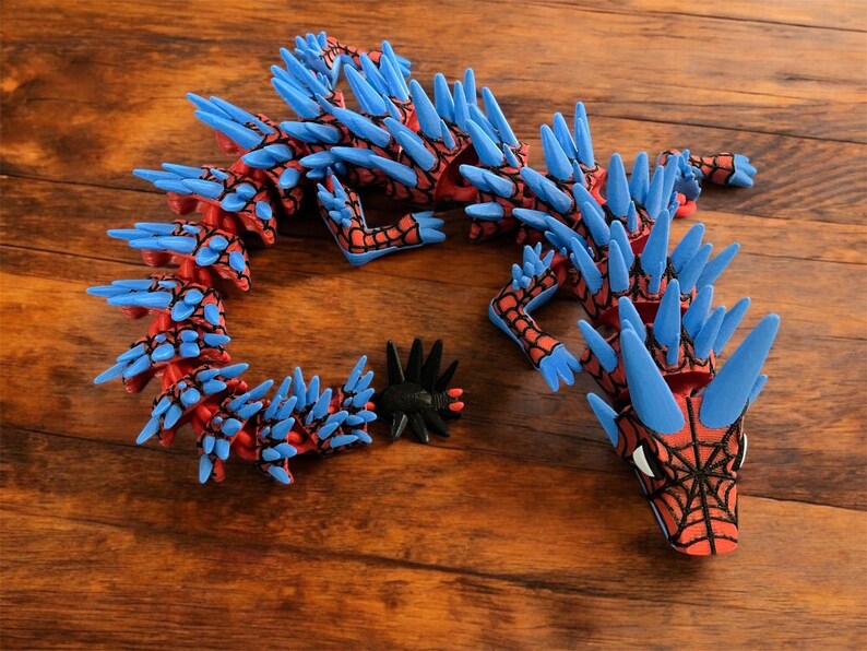 K&ouml;nnte beinhalten: Ein rotes und blaues, bewegliches Drachenspielzeug mit einem Spinnennetzdesign auf dem Kopf. Der Drache hat blaue Stacheln und ist aus Kunststoff. Ein schwarzes, stacheliges Zubeh&ouml;r liegt neben dem Drachen auf einer Holzoberfl&auml;che.