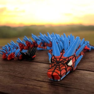 K&ouml;nnte beinhalten: Ein blaues, rotes und schwarzes, bewegliches Drachenspielzeug mit Spinnennetzdesign auf dem Gesicht. Der Drache hat blaue Stacheln entlang seines R&uuml;ckens und Schwanzes. Das Spielzeug liegt auf einer Holzoberfl&auml;che.