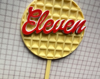 May include: A yellow waffle-shaped cake topper with the word "Eleven" in red and white. The topper has a stick for easy insertion into a cake. The design is reminiscent of a popular television show.