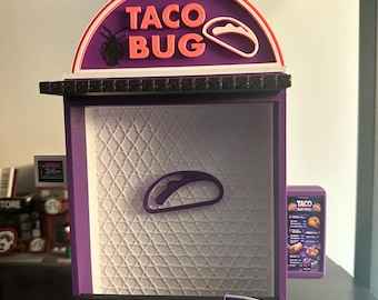 May include: A miniature Taco Bug restaurant model, featuring a purple and white color scheme. The sign reads "TACO BUG" in pink lettering. A small menu and a packet of "HOT BUG SAUCE" are also visible.