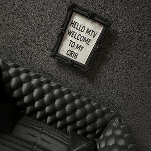 Puede incluir: Un pequeño letrero negro enmarcado con el texto "HELLO MTV WELCOME TO MY CRIB" se encuentra junto a un pequeño sofá negro con un patrón acolchado. El letrero tiene un fondo blanco con texto negro.