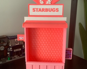 May include: A pink and white 3D printed model of a Starbucks store. The store has a red star on top with two white bugs on it. The sign says "STARBUGS".